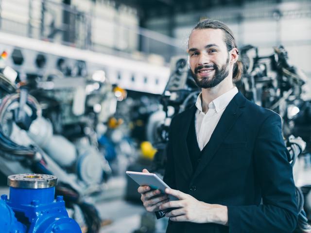 Un hombre sonriente con una tablet en las manos se encuentra en un entorno industrial con máquinas y robots al fondo.