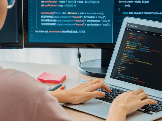 Una persona programando en una laptop frente a dos monitores que muestran código.