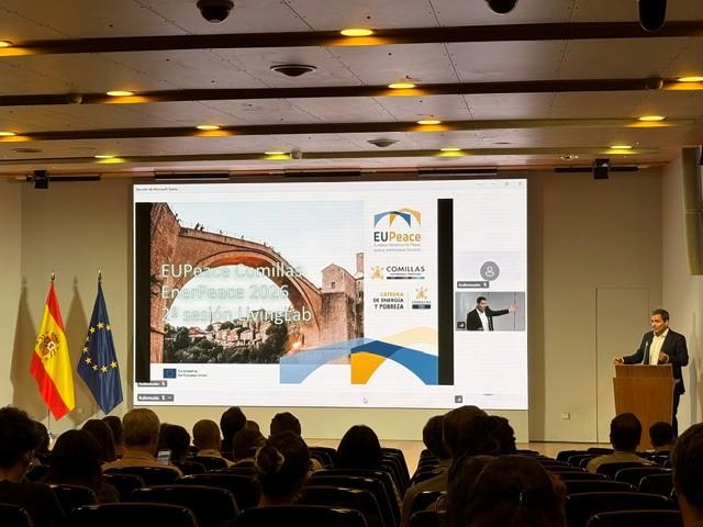 Un conferencista presenta en una sala con banderas de España y la Unión Europea detrás de él, con una proyección de la presentación sobre un evento de paz en pantalla.