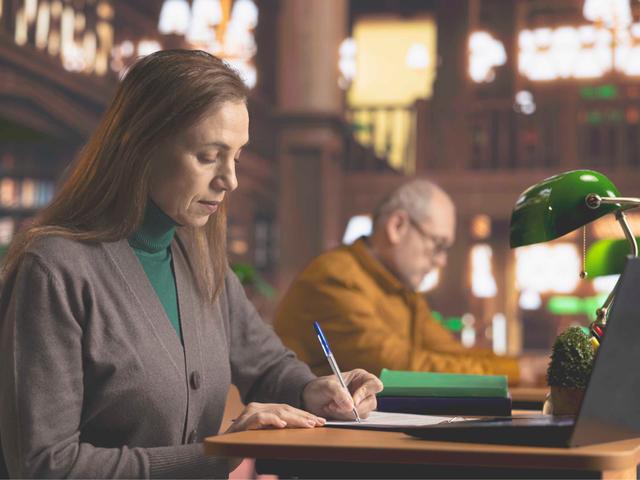Una mujer está escribiendo en una mesa mientras un hombre la observa al fondo.