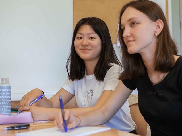 Dos jóvenes sentadas en un aula, sonriendo mientras escriben en sus cuadernos.