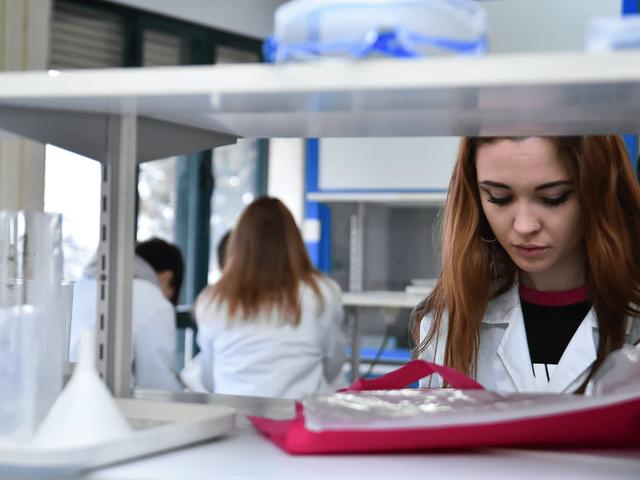 A young woman in a laboratory wearing a white coat examines scientific equipment.