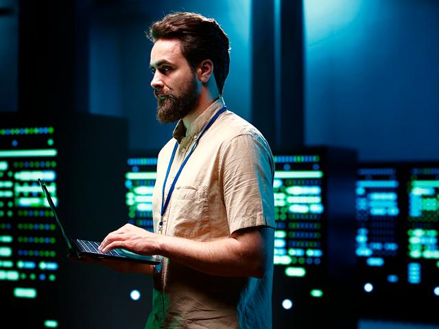 Un hombre con barba sostiene un portátil y se encuentra frente a paneles de datos luminosos en una habitación oscura.
