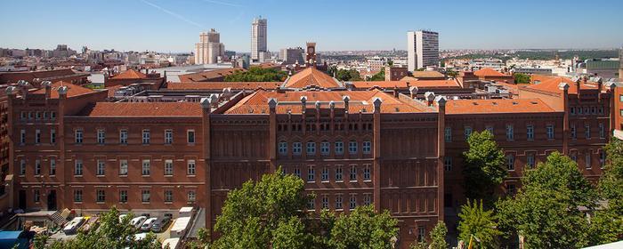 Vista aérea de un gran edificio de ladrillo rojo con múltiples ventanas, rodeado por un paisaje urbano bajo un cielo claro.