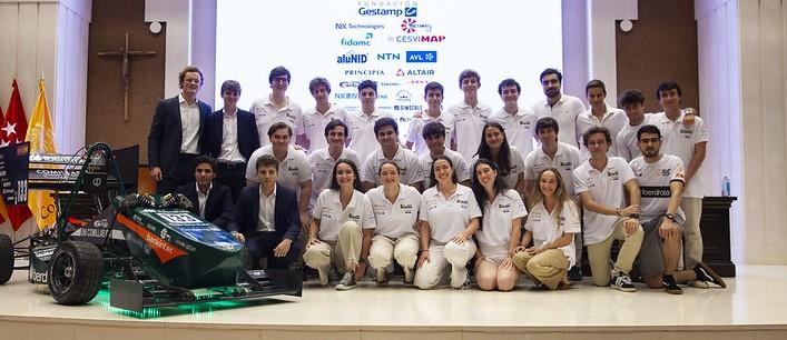 Un grupo de estudiantes posa junto a un coche de carreras en un evento académico.