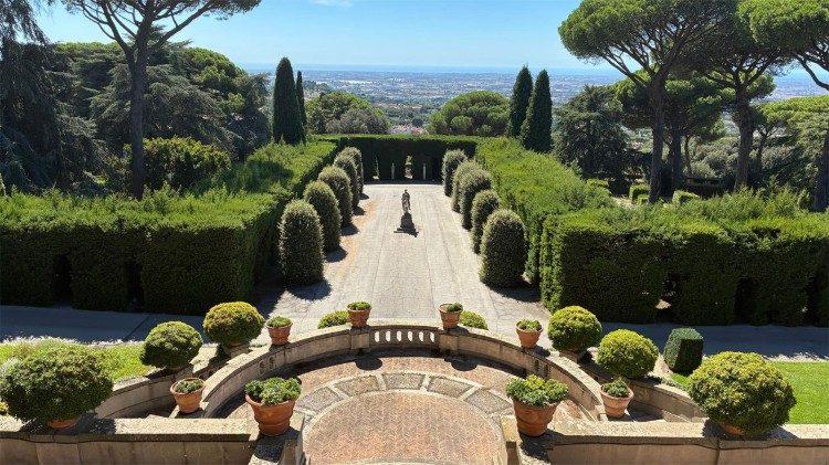 Una vista de un jardín bien cuidado con caminos de piedra y plantas ornamentales, rodeado de árboles altos y un paisaje lejano.