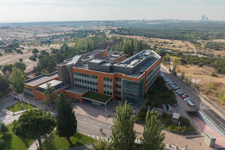 Aerial view of a modern educational building surrounded by green areas and parking lots, with a city skyline in the background.