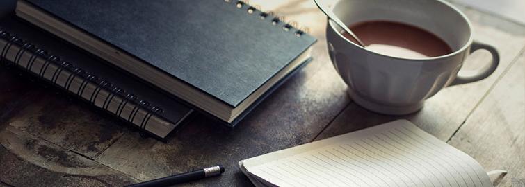 A serene workspace setup featuring an open notebook, a pen, a closed book, and a cup of tea on a wooden surface.