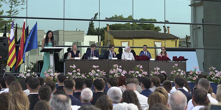 A woman is giving a speech at an outdoor graduation ceremony, with several people seated on stage and an audience in front.