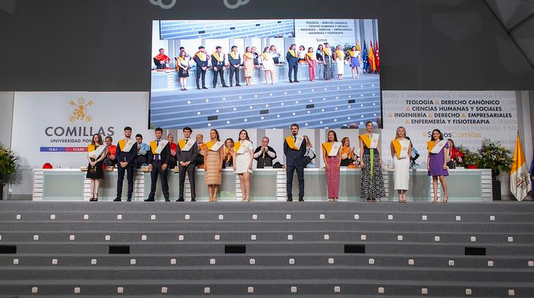 A group of people standing on a stage at a graduation ceremony at Comillas University, with a background sign listing various academic faculties.