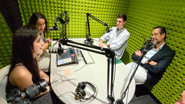 Four people are seated at a round table in a studio, engaged in a podcast recording with microphones and a digital audio mixer.