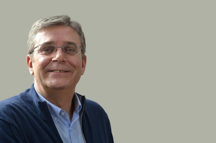 A portrait of a smiling middle-aged man with glasses, wearing a blue sweater over a blue shirt against a light gray background.