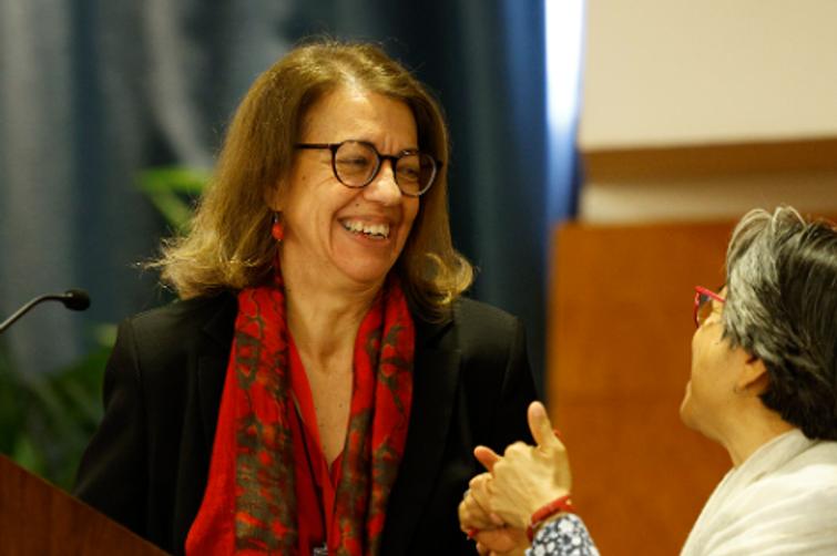 A woman in glasses and a red scarf is laughing while interacting with another person who is gesturing with their hand.