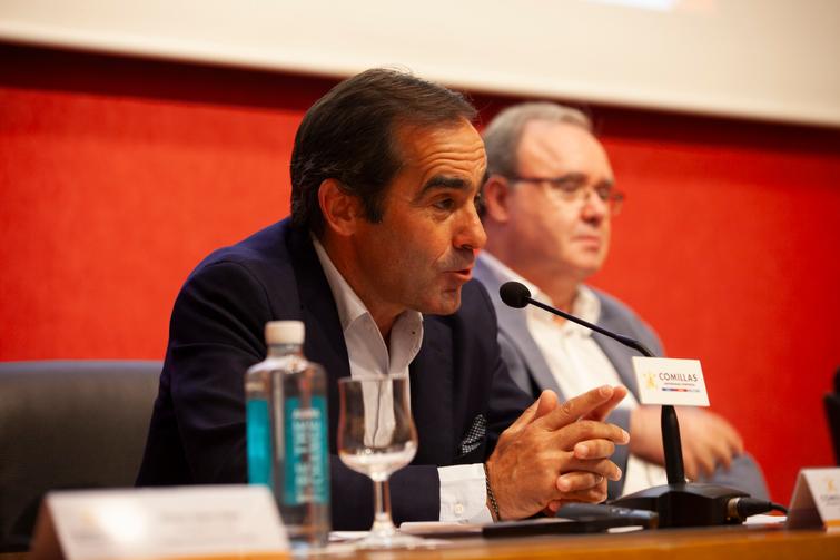 A man in a dark suit speaking into a microphone at a panel discussion, with another man and a water bottle in the background.