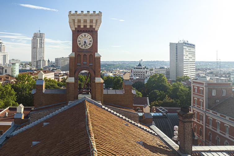 View of a historic tower with a clock, overlooking a cityscape with modern buildings in the background under a clear blue sky.