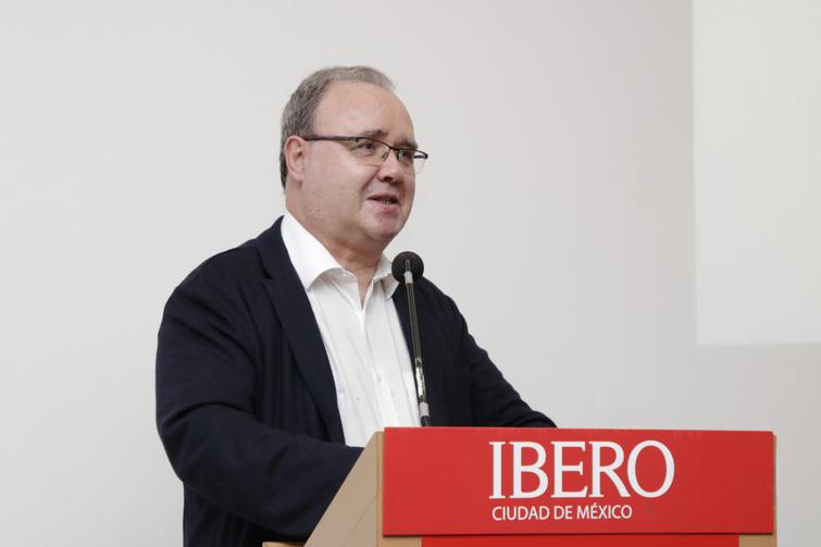 A middle-aged man in glasses speaking at a podium with a sign that reads 'IBERO Ciudad de Mexico'.