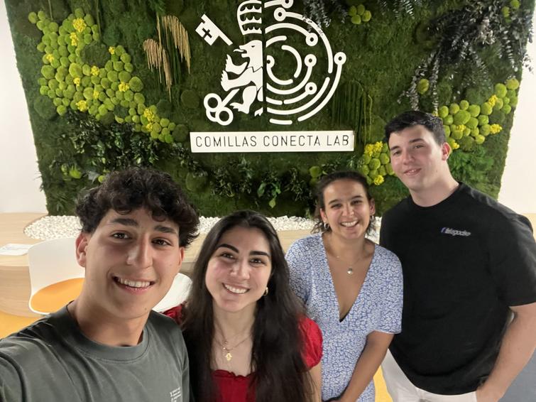 Four young people smiling for a photo in a room with a green botanical wall displaying the 'Comillas Conecta Lab' logo.