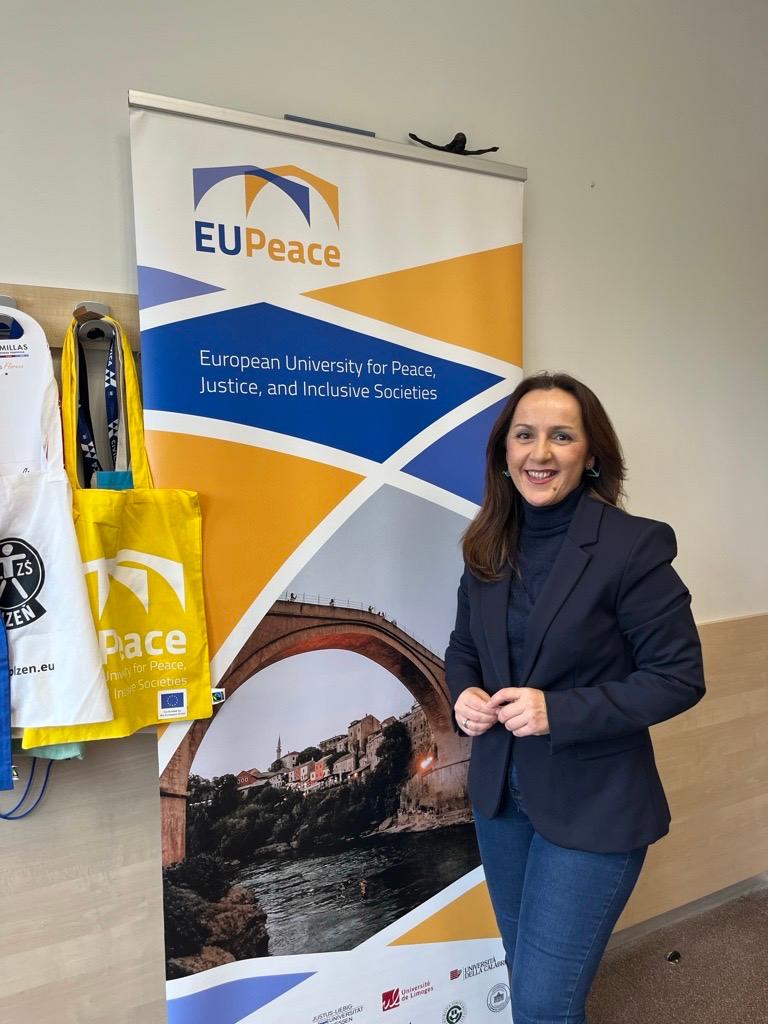 Una mujer sonriente de pie junto a un cartel de la Universidad Europea de la Paz.