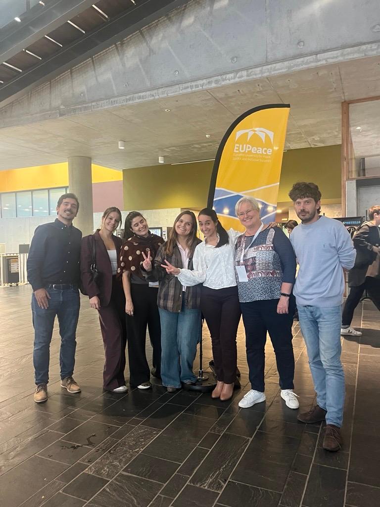 Un grupo de personas sonrientes posando juntas frente a un banner de EUPeace en un ambiente interior moderno.