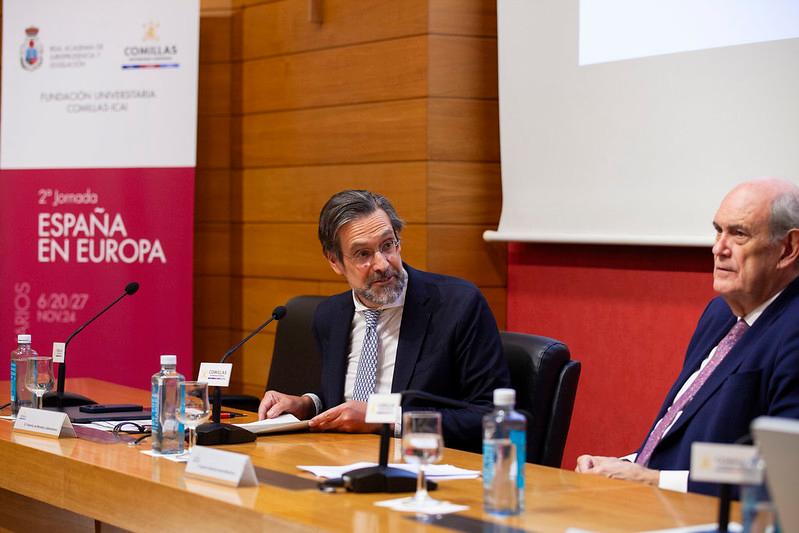 Dos hombres sentados en una mesa durante un evento académico, discutiendo frente a un micrófono con una pancarta que dice 'España en Europa'.