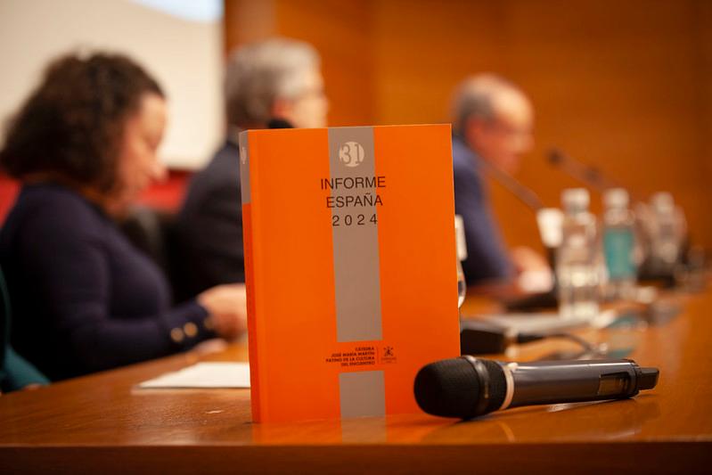 Un libro titulado 'Informe España 2024' situado sobre una mesa durante una conferencia con varios participantes desenfocados al fondo.