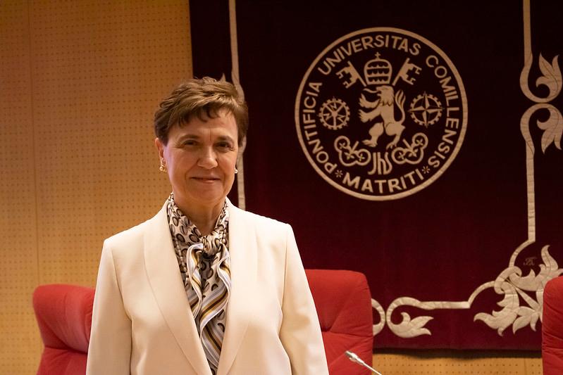 Una mujer de cabello corto vestida con traje blanco y pañuelo al cuello posa delante de un escudo universitario.