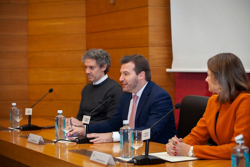 Tres personas sentadas en una mesa durante un evento formal, hablando entre ellas y con micrófonos delante.