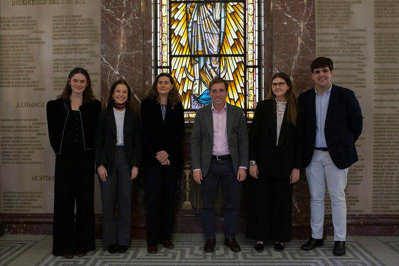 Grupo de seis personas posando frente a una ventana de vitral en un entorno académico.
