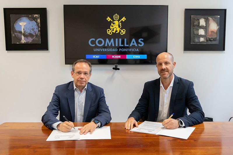 Dos hombres firmando documentos en una mesa frente a un televisor con el logo de la Universidad Pontificia Comillas.