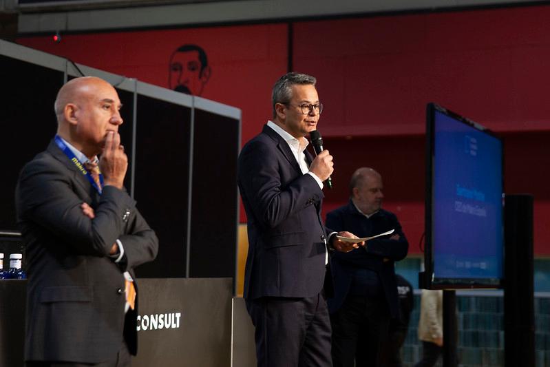 Un hombre habla frente a un micrófono en un evento mientras otro lo observa en segundo plano.