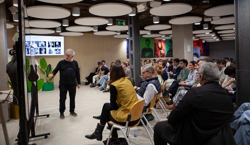 Una conferencia en un espacio moderno con una audiencia atenta frente a un presentador.