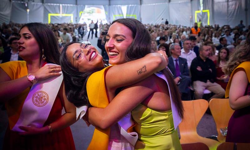 Dos jóvenes se abrazan con alegría en un evento de graduación mientras el público celebra al fondo.
