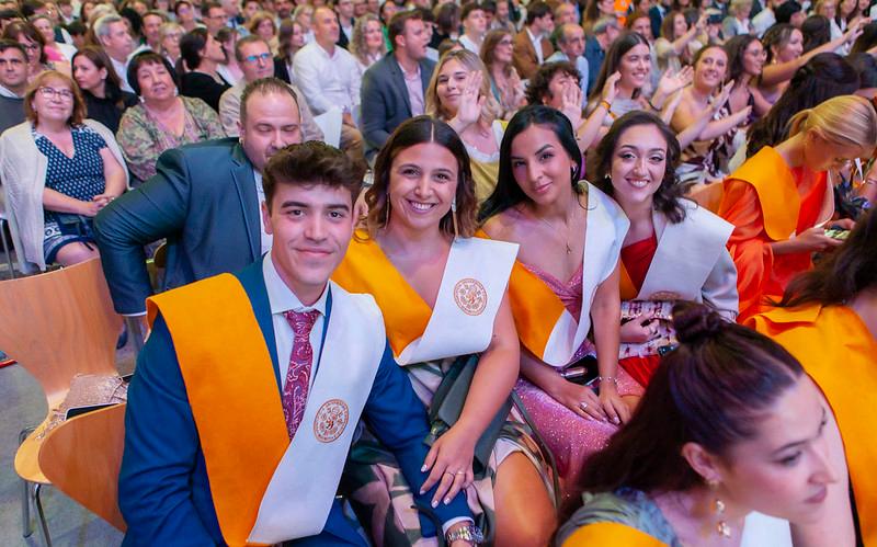 A group of young graduates seated in an auditorium, donning ceremonial robes with orange sashes, smiling at a graduation ceremony.