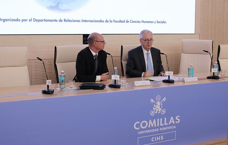 Dos hombres sentados frente a un estrado en una conferencia en la Universidad Pontificia Comillas.