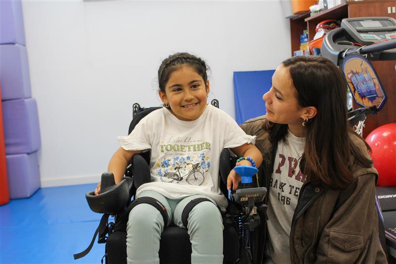 Una niña sonriente en una silla de ruedas es asistida por una joven en un entorno de terapia.