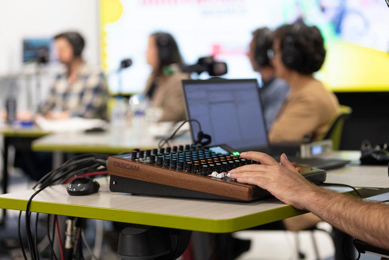 Un grupo de personas sentadas en una mesa, participando en una grabación o transmisión, mientras un individuo controla un equipo de audio.