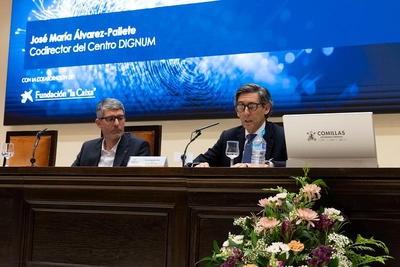 Dos hombres están sentados en la mesa de un evento, uno de ellos hablando mientras el otro escucha.