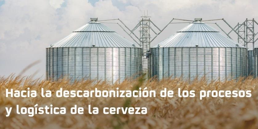 La imagen muestra silos metálicos en un campo de trigo bajo un cielo nublado, acompañados de un mensaje sobre la descarbonización en la logística de la cerveza.
