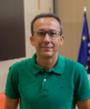 A middle-aged man wearing glasses and a green polo shirt smiling in front of a blurred European Union flag.