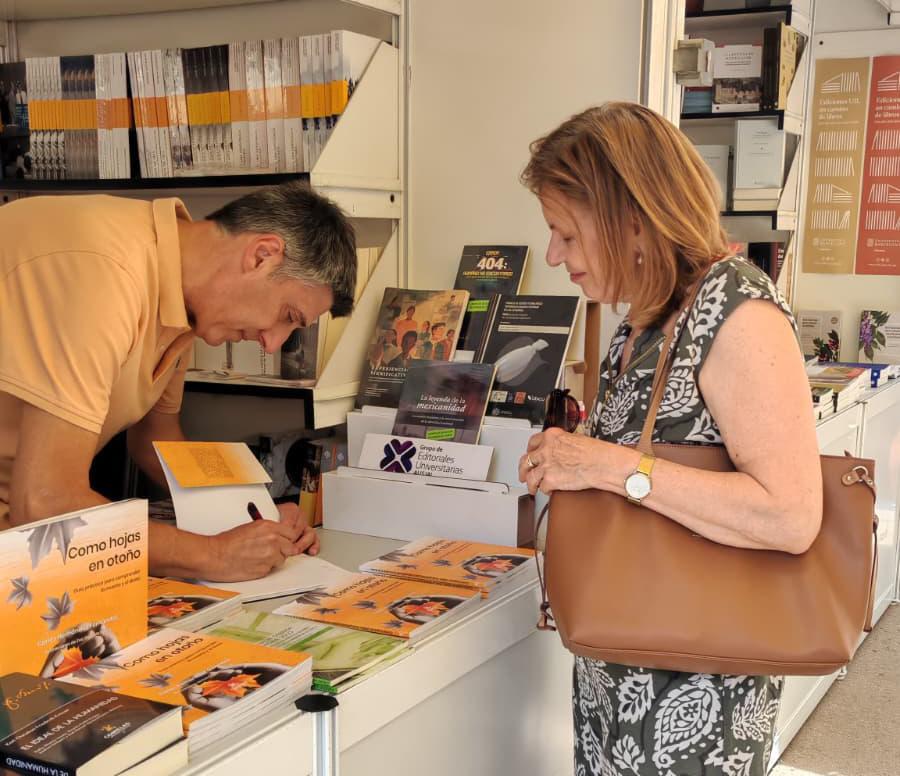 Un hombre firma un libro mientras una mujer lo observa en un evento literario.