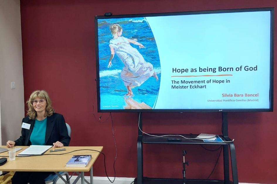 Una mujer sonriente se encuentra sentada frente a una presentación en pantalla sobre la esperanza en Meister Eckhart.