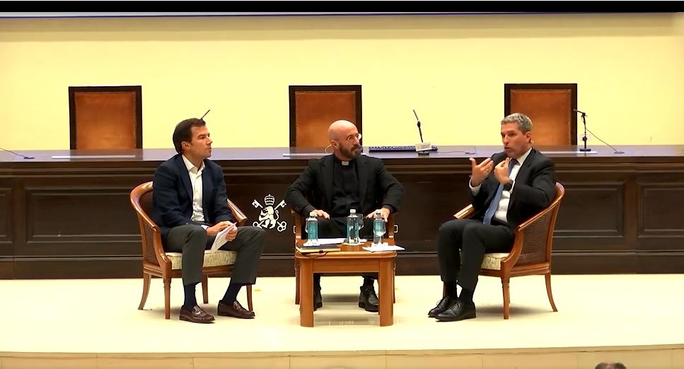 Tres hombres sentados en sillas de madera en un escenario, dialogando en un evento formal.