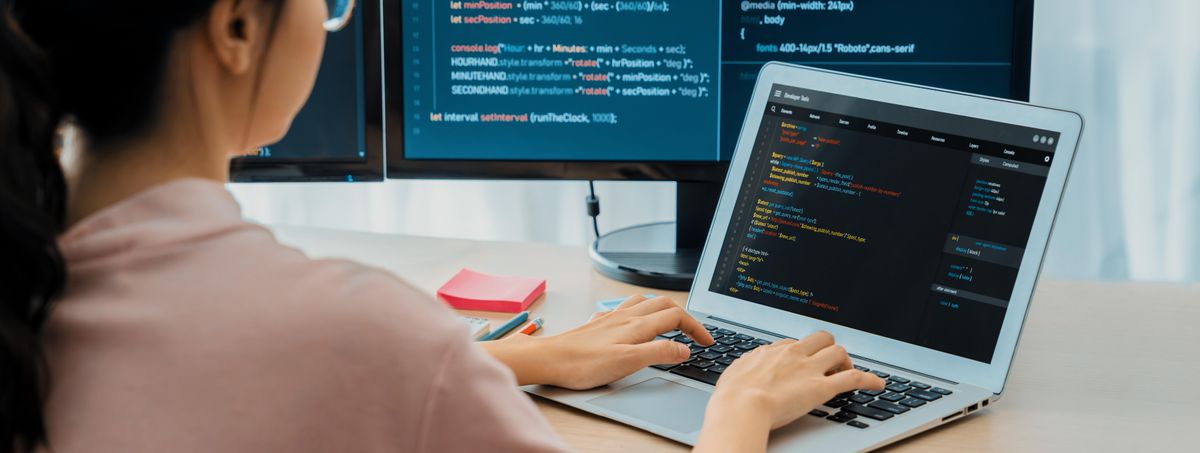 Una persona programando en una laptop frente a dos monitores que muestran código.