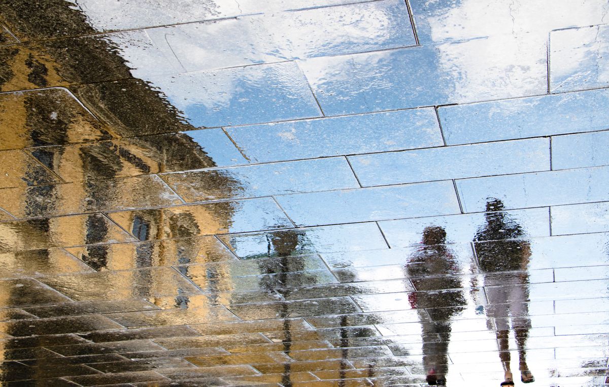 Reflection of two people walking and a building observed on a wet pavement.