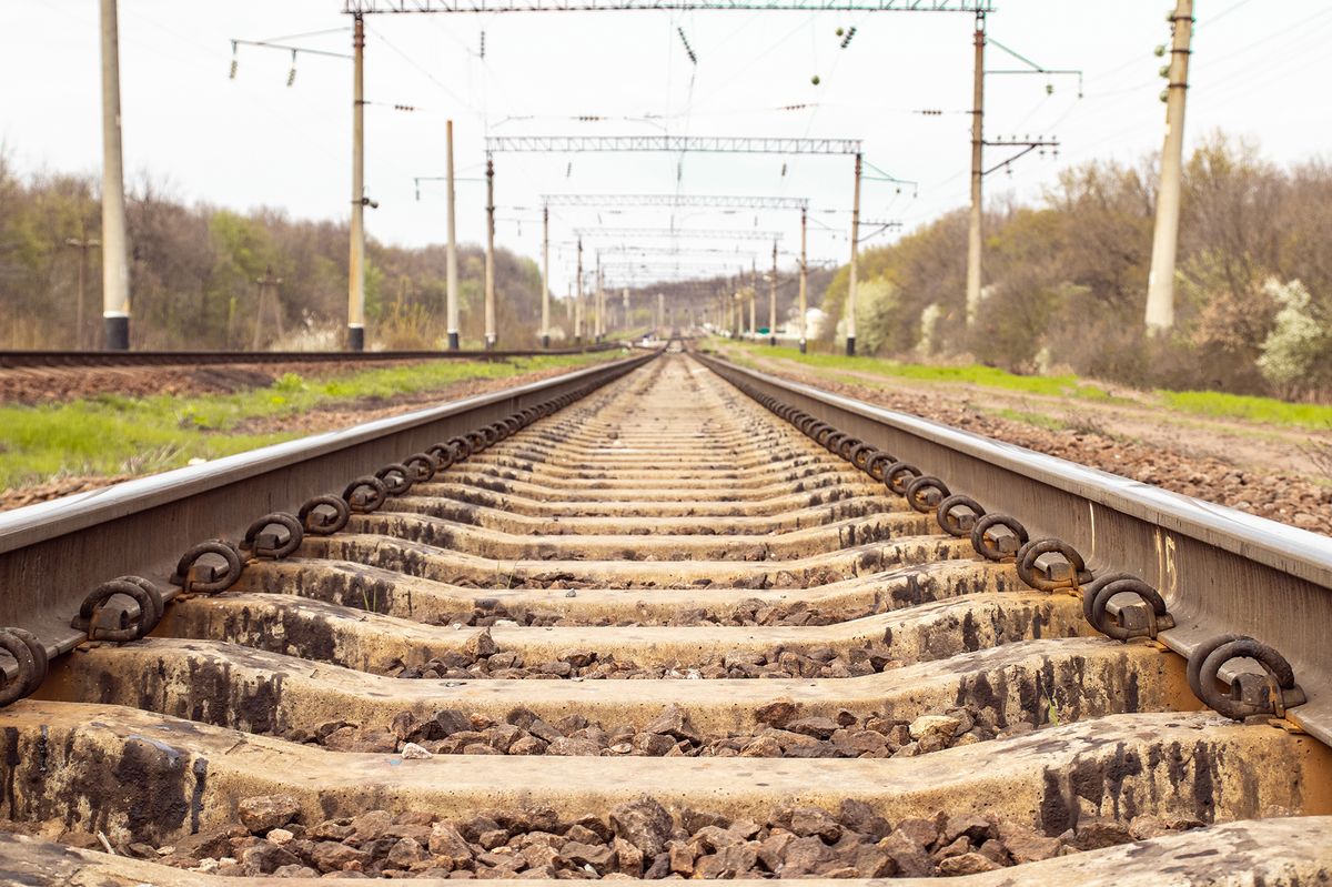 A perspective view of railway tracks receding into the distance with overhead electrical lines and greenery on either side.