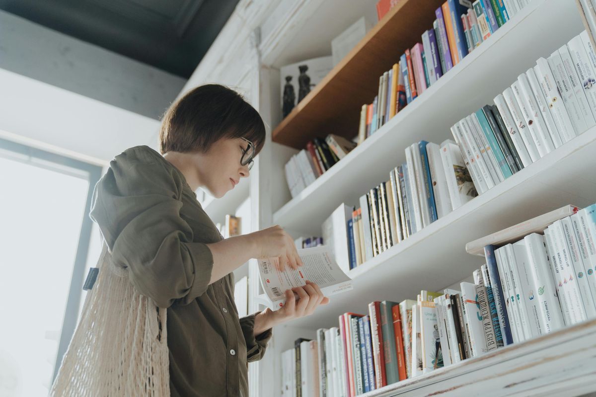 A young woman is browsing books in a well-lit library, standing next to a shelf filled with a variety of books.