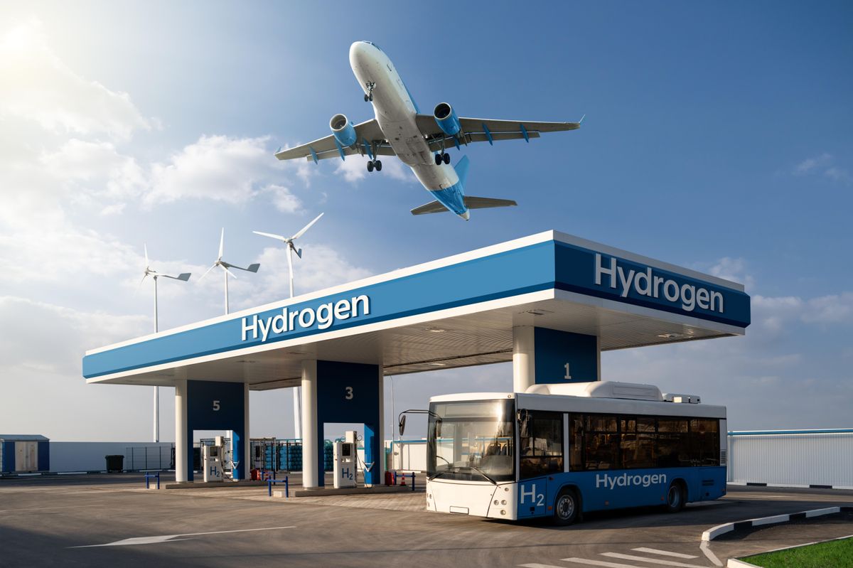 An airplane flies over a hydrogen fuel station servicing a bus, with wind turbines in the background.