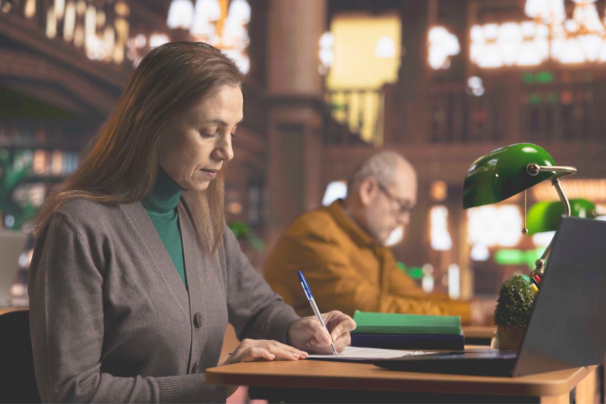 Una mujer está escribiendo en una mesa mientras un hombre la observa al fondo.