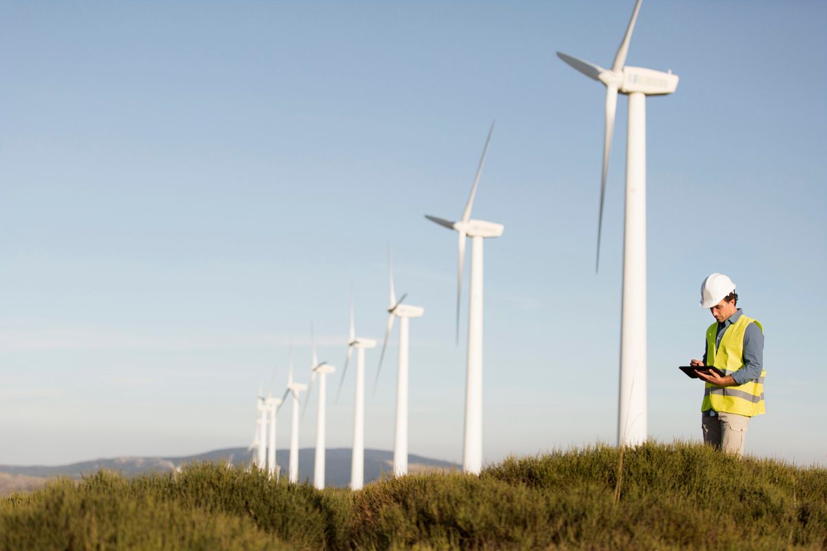 Un técnico revisa un dispositivo mientras se encuentra rodeado de aerogeneradores en un paisaje abierto.