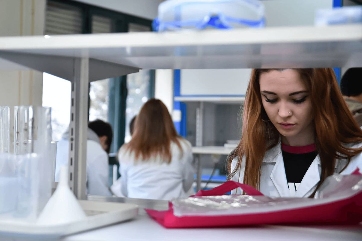A young woman in a laboratory wearing a white coat examines scientific equipment.